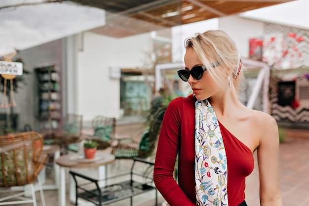 Attractive joyful lady with blond hair wearing cherry sweater with bare shoulder black sunglasses and modern accessory in the head is looking away against entertainment district