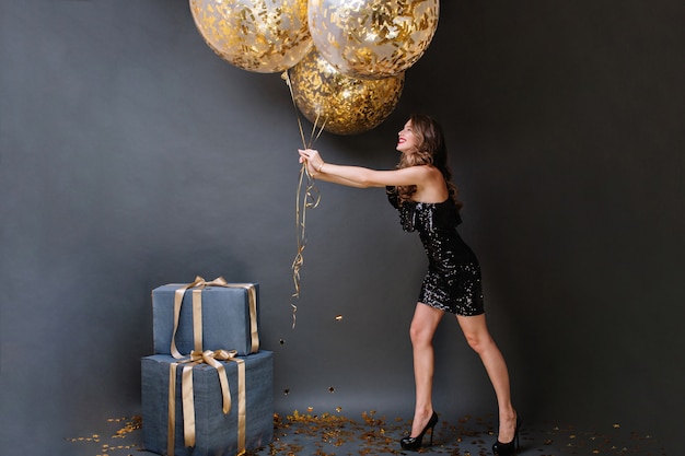 Attractive joyful young woman in black luxury dress having fun with big balloons full with golden tinsels. Happy birthday party, presents, smiling, expressing positivity.