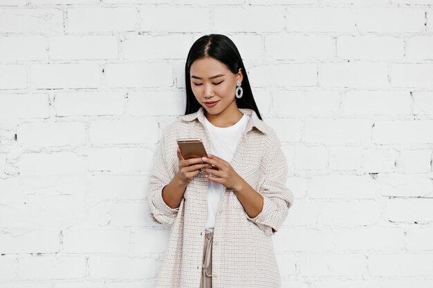 Attractive lady in beige cardigan and t-shirt chats in phone on white brick wall wall