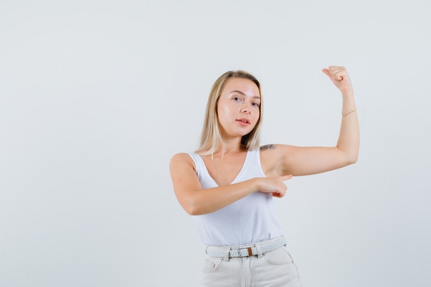 Attractive lady pointing at her arm muscles in white blouse and looking assured