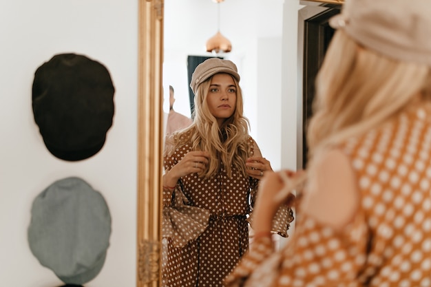 매력적인 아가씨는 골드 프레임에 거울 앞에서 Preens. 세련 된 모자에 곱슬 여자는 밝은 아파트에서 포즈.