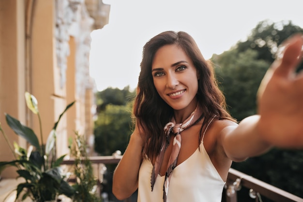 Attractive lady in silk white top takes selfie on balcony. Pretty brunette woman in beige shawl makes photo in sunshine.