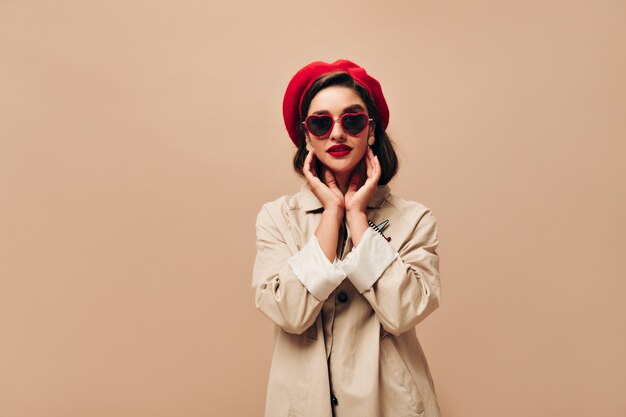 Attractive lady in sunglasses and red beret poses on beige background. Wonderful girl in autumn light coat and bright hat looks at camera.