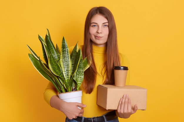 Attractive lady with long straight hair wearing yellow shirt,  with contemptuous facial expression, holding carton box, take away coffee and flower in hands.
