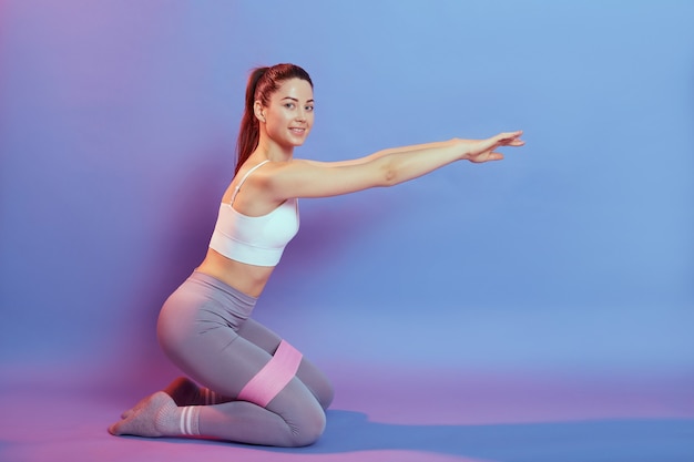 Attractive lady with ponytail training inner thighs with rubber elastic resistance, looks at camera with smile, training body, dresses sportswear, isolated over color background.