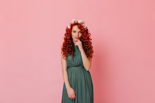 Attractive lady with red curls and blue eyes looking at camera. Woman in long green dress and roses in her hair posing on pink space.