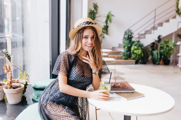 Attractive long-haired girl resting in restaurant and sitting at the table with magazines touching chin with fingers