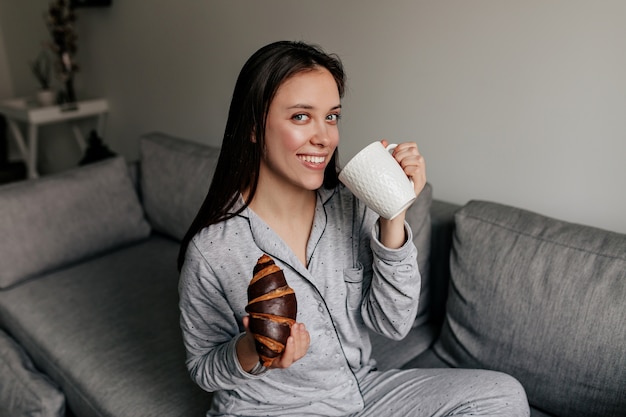 晴れた日にコーヒーを飲みながら家でクロワッサンを食べてパジャマを着て素晴らしい笑顔で魅力的な素敵な女性。