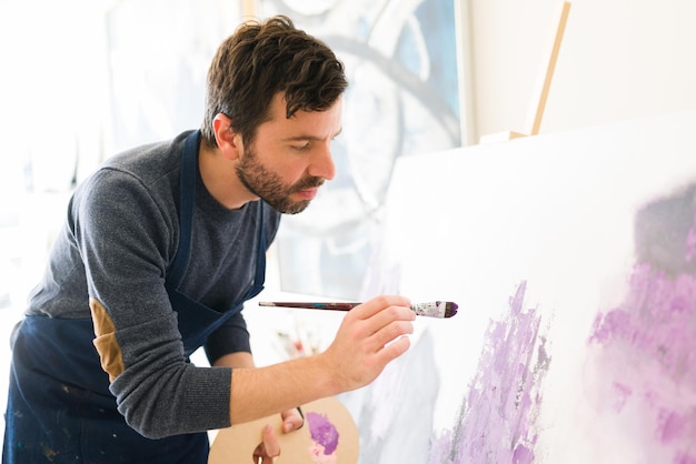Attraente artista maschio sulla trentina sta dando gli ultimi tocchi di pittura al nuovo dipinto colorato che sta facendo nel suo studio d'arte