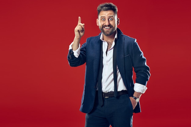 Attractive male half-length front portrait on red  wall. Young emotional surprised bearded man presenting something. Human emotions, facial expression concept.