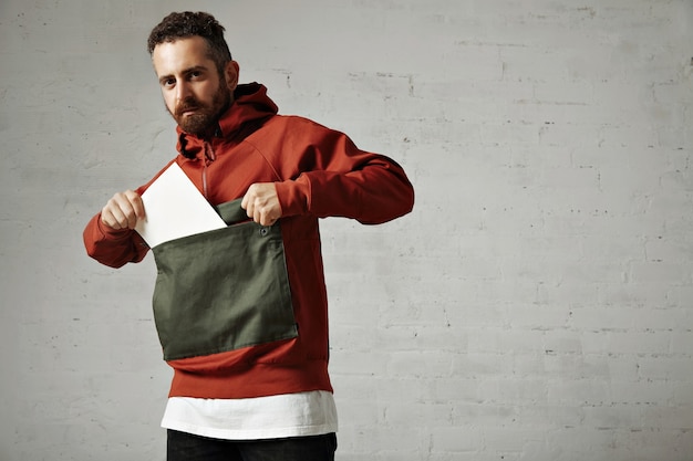 Attractive male model taking a blank white sheet of paper out of the front pocket of his red and gray parka on white