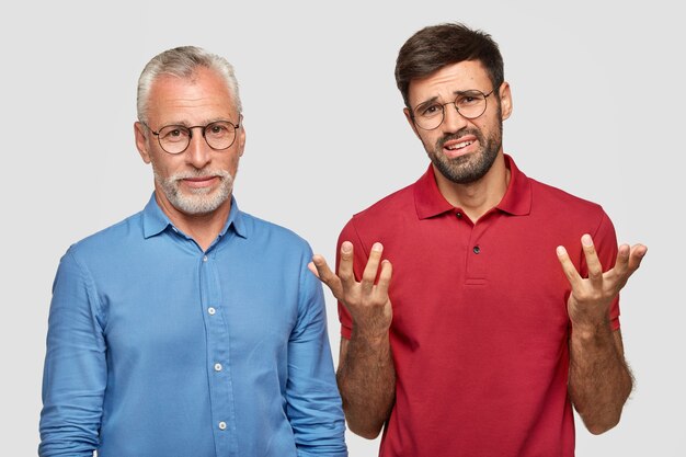 Attractive male pensioner cooperates together with his young colleague who has desperate nervous expression, stand next to each other, isolated over white wall. People and relationships