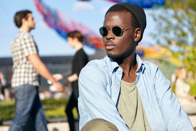 Free photo attractive male with dark pure healthy skin and bristle, wearing shades and shirt, looking seriously aside while sitting outdoors