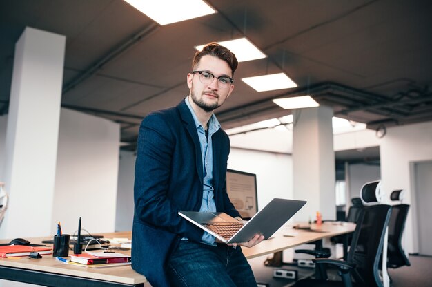 Attractive man in glassess is sitting near the workplace in office. He wears  blue shirt, dark jacket.  He holds laptop and looks to the camera.