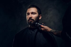 attractive man have beard trimming with machine at dark barbershop from female.