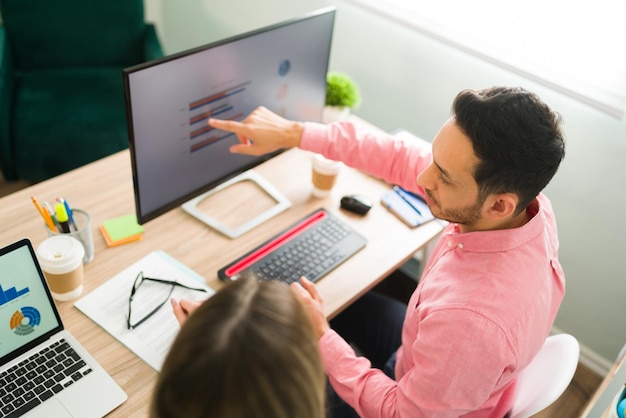 Attractive man pointing to the computer screen and business presentation while sitting at the office. Professional coworkers working on an analytical report