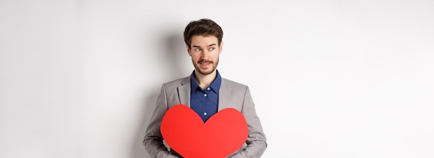 Attractive man in suit looking left and smiling holding red heart cutout prepare valentines day surp