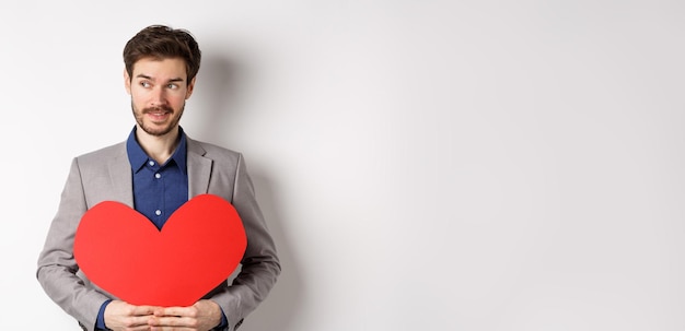 L'uomo attraente in giacca e cravatta che guarda a sinistra e sorride tenendo il ritaglio del cuore rosso prepara il surp di san valentino