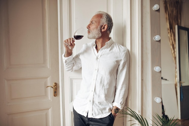 Attractive man in white shirt drinking wine in apartment Pensive guy in classic suit posing with glass of red drink in his hands
