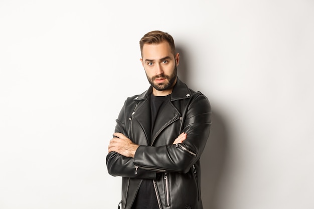 Attractive  man with beard, wearing leather jacket smiling, looking confident with hands crossed on chest 