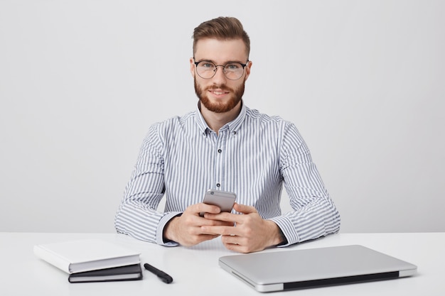 attractive man with fashionable hairstyle and thick reddish beard, wears rounded glasses and formal shirt,