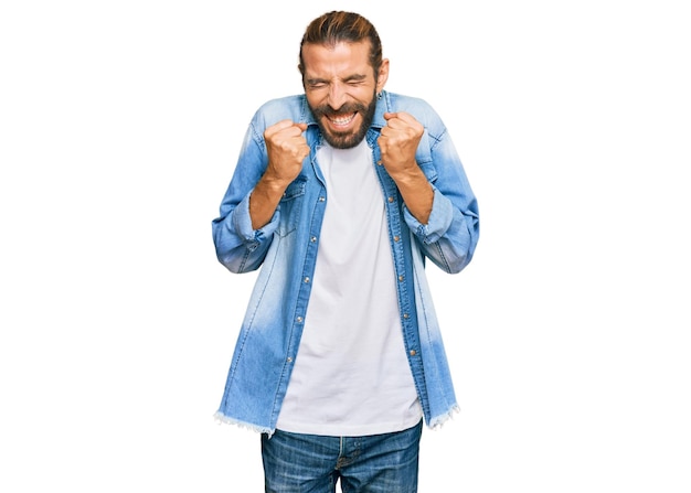 Attractive man with long hair and beard wearing casual denim jacket excited for success with arms raised and eyes closed celebrating victory smiling winner concept