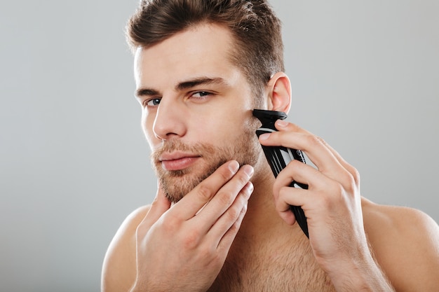 Attractive masculine male being undressed at home having skincare while shaving his face with trimmer against grey wall