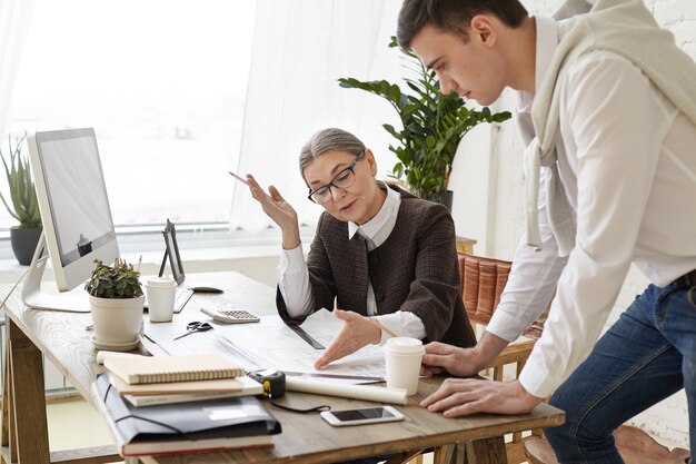 Attractive mature female architect in glasses sitting in front of computer and checking technical drawings by her trainee, indicating drawbacks, sharing her ideas and vision. Job and cooperation