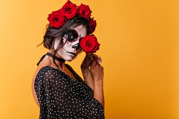 Attractive mature woman in Halloween outfit loves roses. Closeup portrait of mexican woman closing her eyes with red flower.