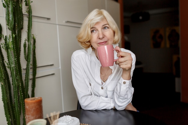 매력적인 중년 금발의 여자가 부엌에서 집에서 휴식과 커피를 마시는