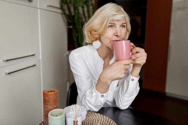 Привлекательная блондинка средних лет отдыхает дома на кухне и пьет кофе