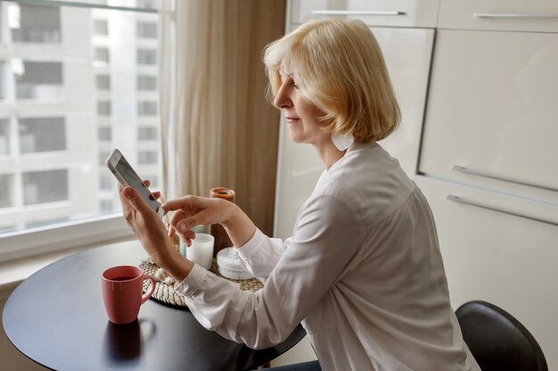 Attractive middle-aged blonde woman relaxing at home on the kitchen and talking by mobile phone