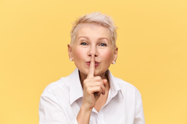Attractive middle aged female in white shirt holding index finger at her mouth, making shush sign, saying Shh, donât tell anyone, asking you to keep her secret, having mysterious facial expression