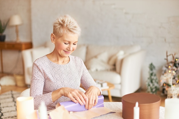 Attractive middle aged female with blonde short haircut sitting at table in living room, wrapping Christmas present for husband in gift paper