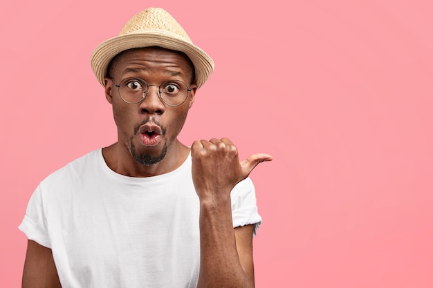 Attractive middle aged man agricultural worker in casual clothes and hat, indicates aside with thumb, shows blank space against pink wall