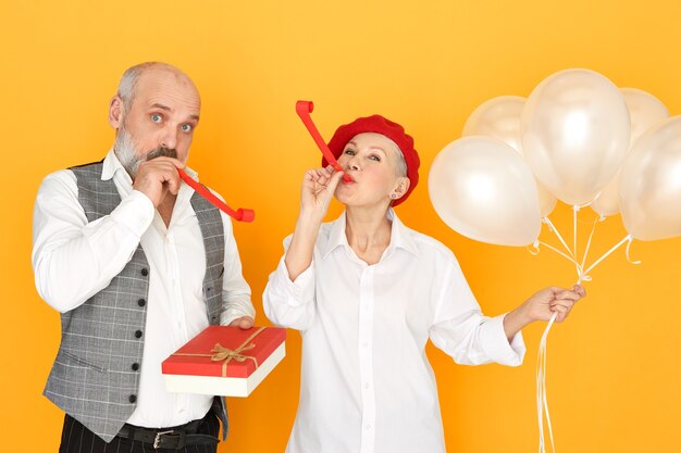 Attractive middle aged woman posing isolated with helium balloons with bearded senior male holding box of chocolate, blowing whistles