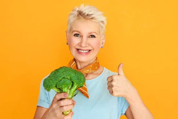 신선한 브로콜리를 들고 건강 한 유기농 식품을 선택하는 카메라에 웃 고 짧은 요정 머리를 가진 매력적인 중간 나이 든된 여자.
