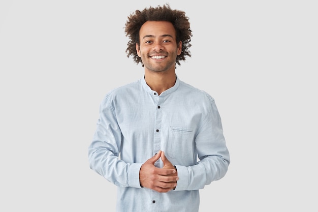 Attractive mixed race male with positive smile, shows white teeth, keeps hands on stomach, being in high spirit, wears white shirt, rejoices positive moments in life. People and emotions concept