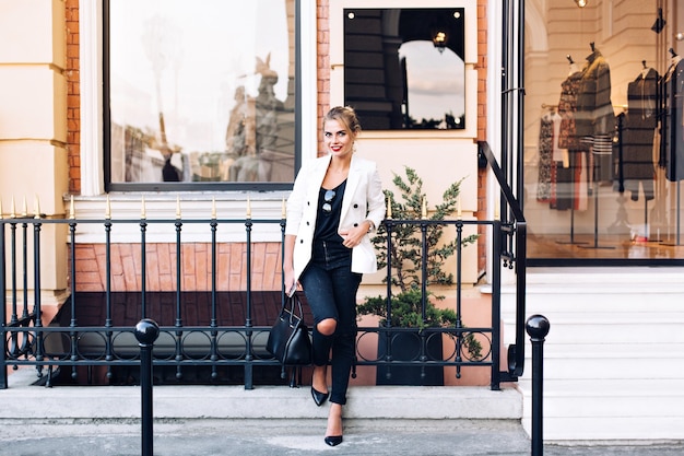 Attractive model in white jacket on heels is leaning on fence on shop background. She keeps hand in pocket, smiling to camera.