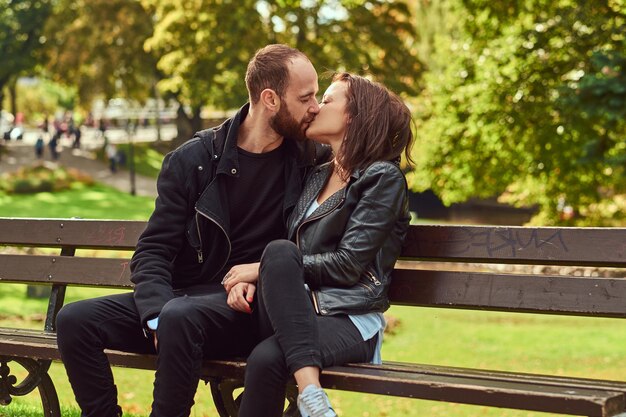 Attractive modern couple kissing on a date while cuddling on a bench in the park. Enjoying their love and nature.