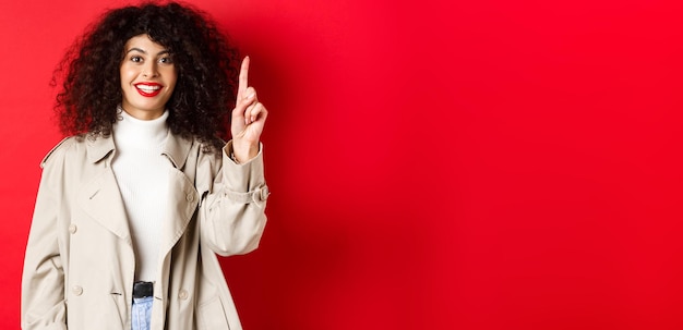 Attractive modern woman with red lips curly hairstyle wearing spring trench coat pointing finger up