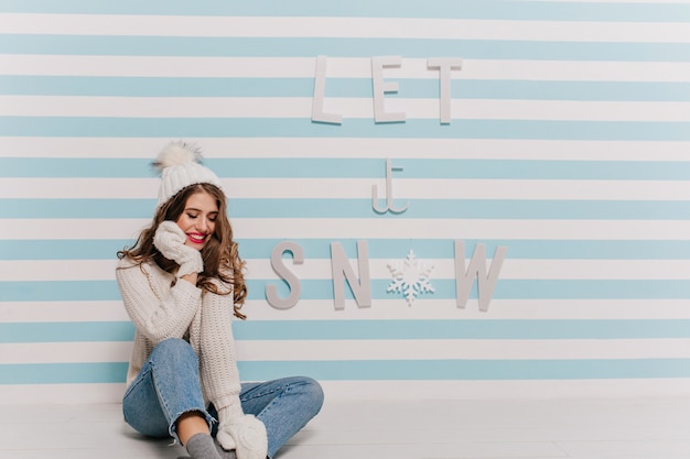 Attractive, modest lady in white knit outfit shyly looks down and touches her beautiful face. portrait with new year lettering on striped wall