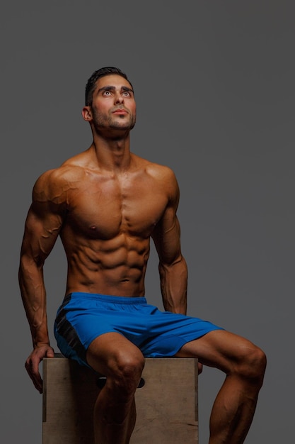 Привлекательный мускулистый парень сидит на подиуме на сером фоне