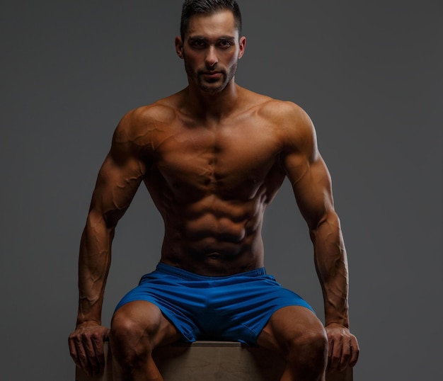 Free photo attractive muscular guy sitting on podium on grey background