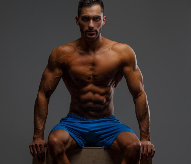 Attractive muscular guy sitting on podium on grey background