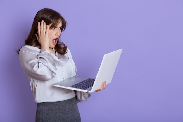 Attractive nervous young business woman with laptop in hands, looking at screen of device with shocked expression, touching her head with hand, keeps mouth widely opened, stands against lilac wall.