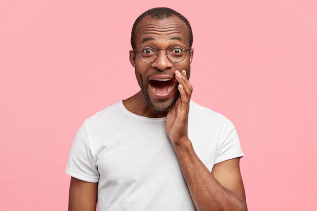 Attractive overjoyed young man expresses happy emotions, enjoys life, exclaims loudly keeps mouth widely opened, dressed in white t shirt, stands against pink wall