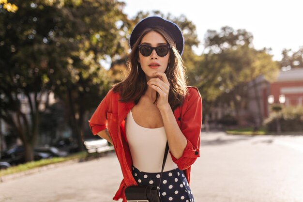 Attraente ragazza pallida in berretto francese occhiali da sole neri top bianco e camicia in posa all'aperto e toccando il viso tempo caldo e soleggiato