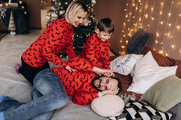 Бесплатное фото Привлекательные родители и их маленький сын в красных свитерах развлекаются, лежа на кровати перед рождеством