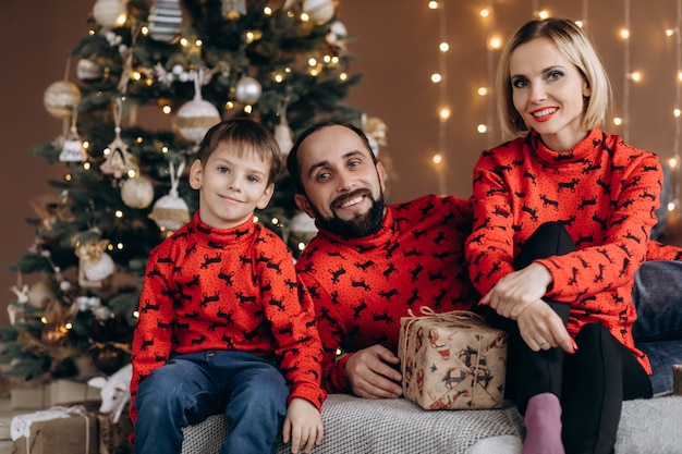 Привлекательные родители и их маленький сын в красных свитерах развлекаются, открывая подарки перед Рождеством